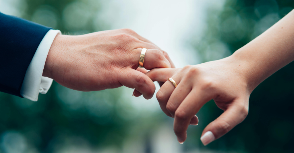 An image of two hands interconnected with just the pinky fingers and wearing soulmate wedding rings.